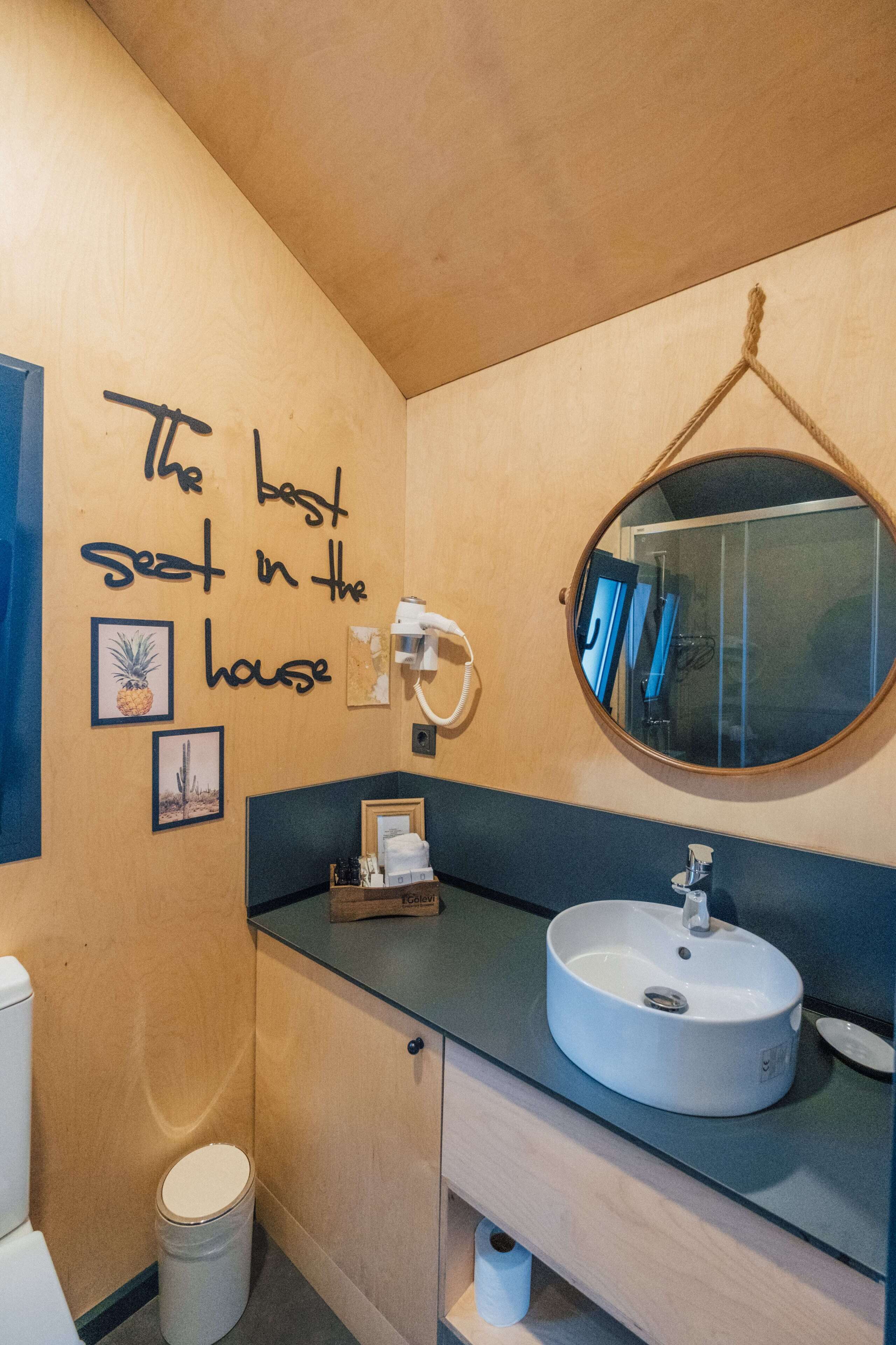 A bathroom with a wooden vanity, a round mirror, a white sink, and a toilet. There is a sign on the wall that reads "The best seat in the house."