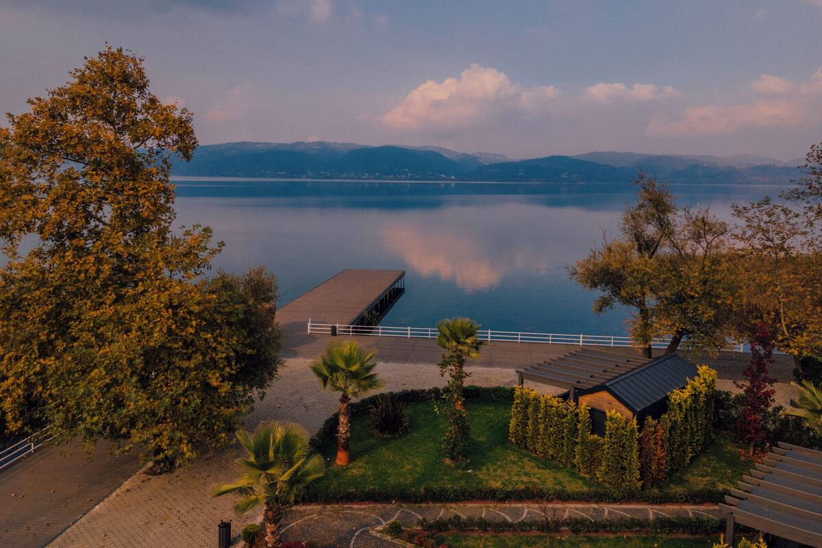 A lakeside view with a wooden deck, a small building with a sloped roof, and a distant mountain range.
