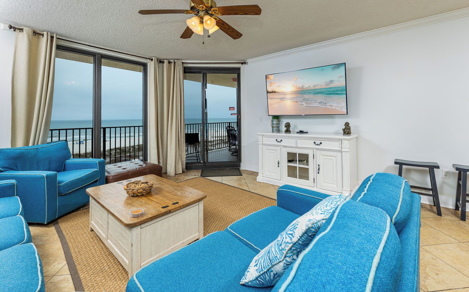 Ein Zimmer mit großem Fenster mit Blick auf den Strand, einem Deckenventilator, einem Holztisch und einer weißen Kommode mit Dekoration.