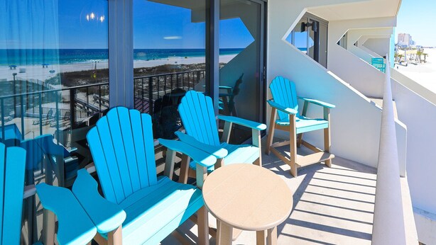 Ein Balkon mit blauen Adirondack-Stühlen und einem kleinen runden Tisch, der auf einen Strand und das Meer hinausblickt.