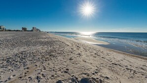 Ein weiter Sandstrand mit Fußspuren, ein klarer Himmel und Gebäude im Hintergrund.