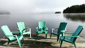 Eine hölzerne Anlegestelle mit vier grünen Adirondack-Stühlen, die auf einen stillen See zeigen, umgeben von Bäumen und einer nebligen Insel im Hintergrund.
