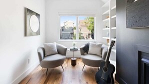 Una sala de estar moderna con dos sillones, una mesa de centro, una guitarra, una chimenea y un ventanal con vistas a casas.