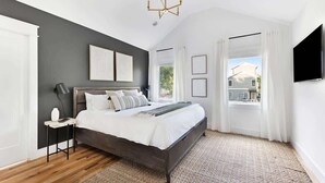 Un dormitorio con cama de madera oscura, ropa de cama blanca, una mesita de noche pequeña, televisor montado en la pared y un ventanal con cortinas.