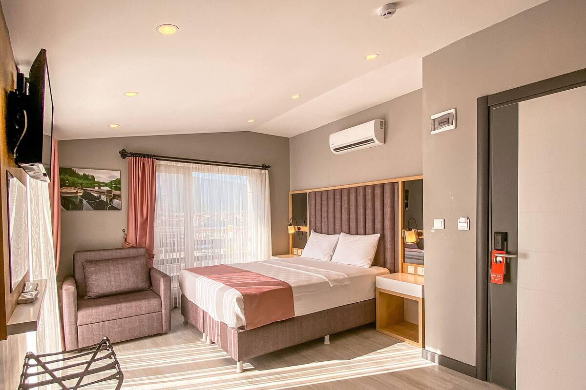 A modern hotel room with a large bed, a seating area, a flat-screen TV, and a window with curtains.