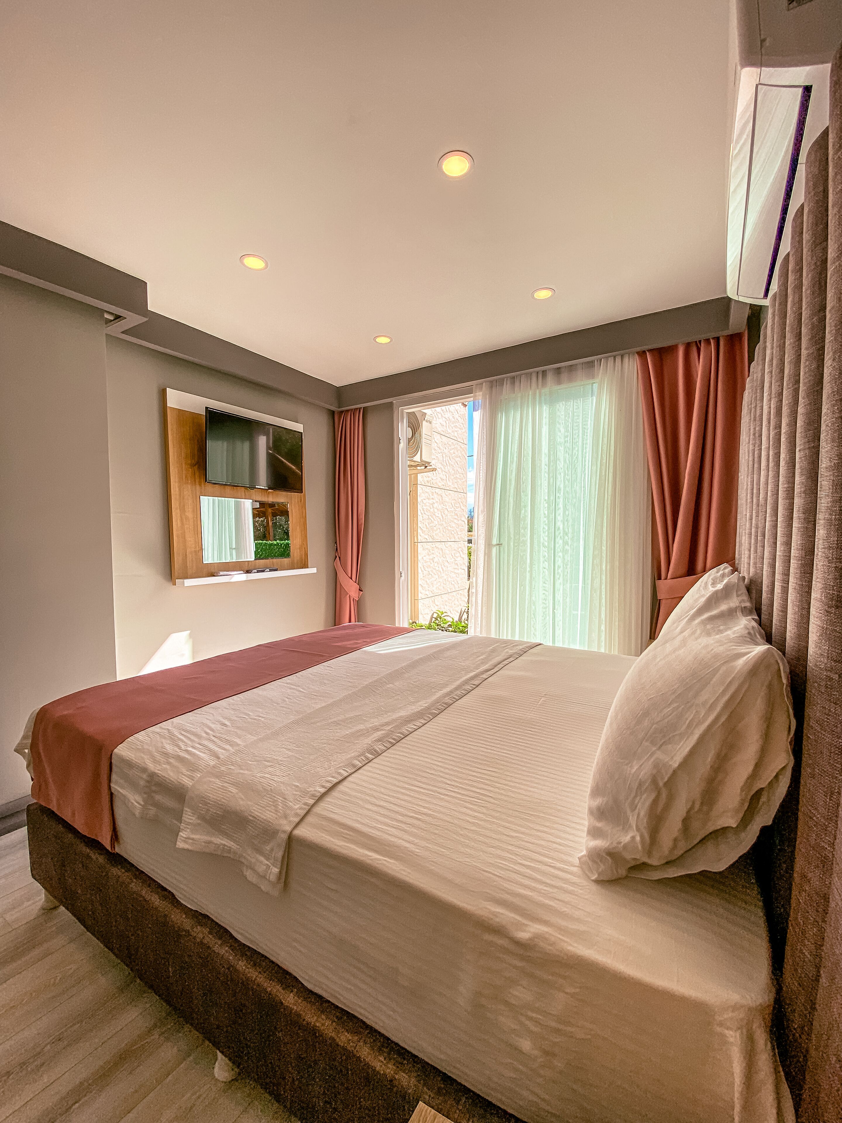 A bedroom with a large bed, a flat-screen TV mounted on the wall, and a window with curtains.