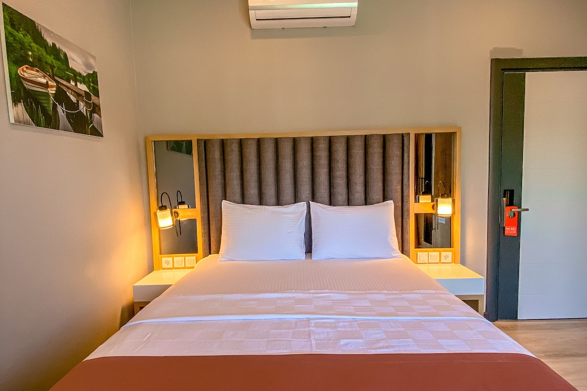 A hotel room with a large bed, a headboard with vertical slats, two bedside tables with lamps, an air conditioning unit, and a door with a red lock.