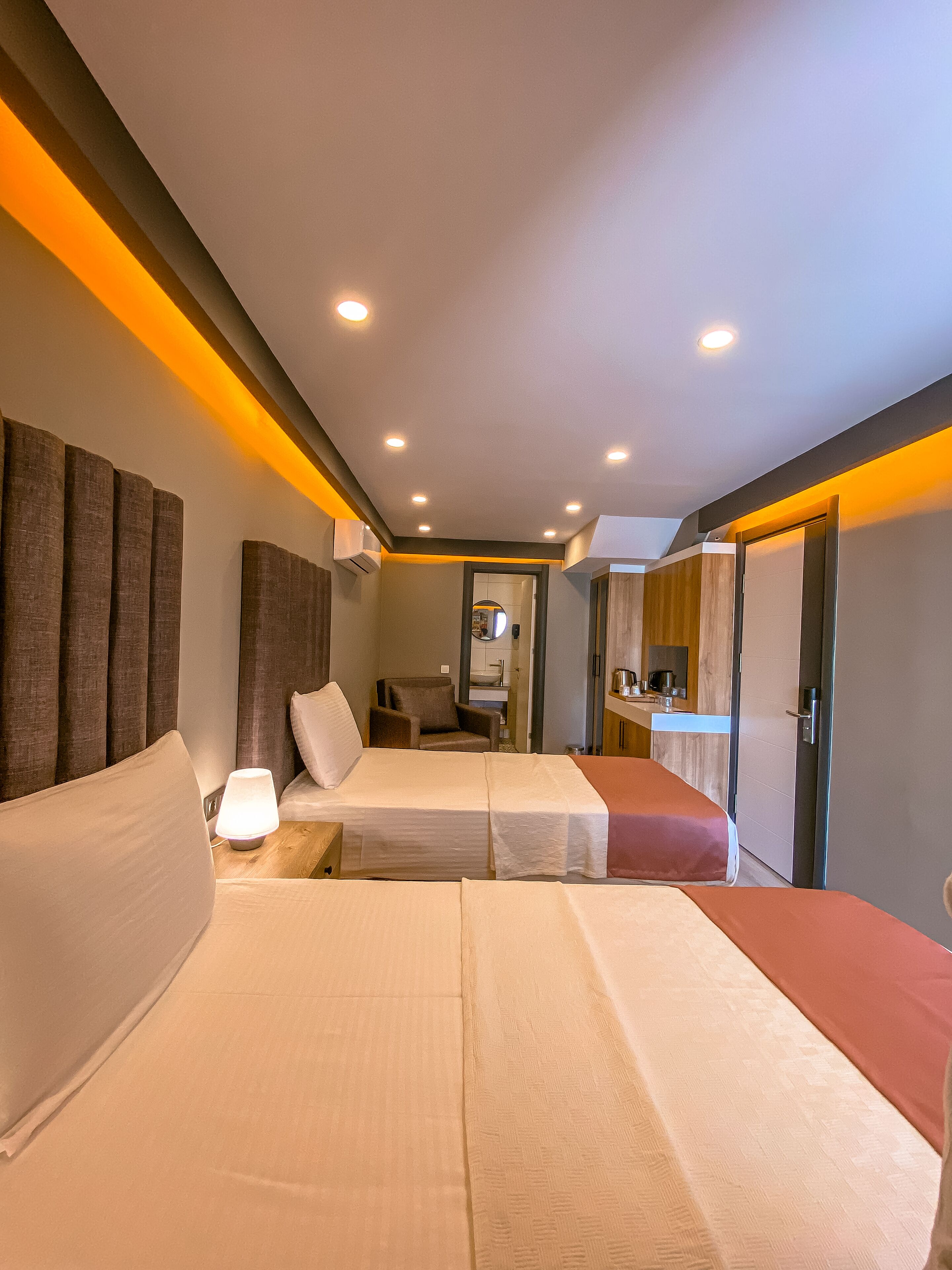 A hotel room with two beds, a bedside table with a lamp, a chair, a small kitchenette, and a bathroom visible through an open door.