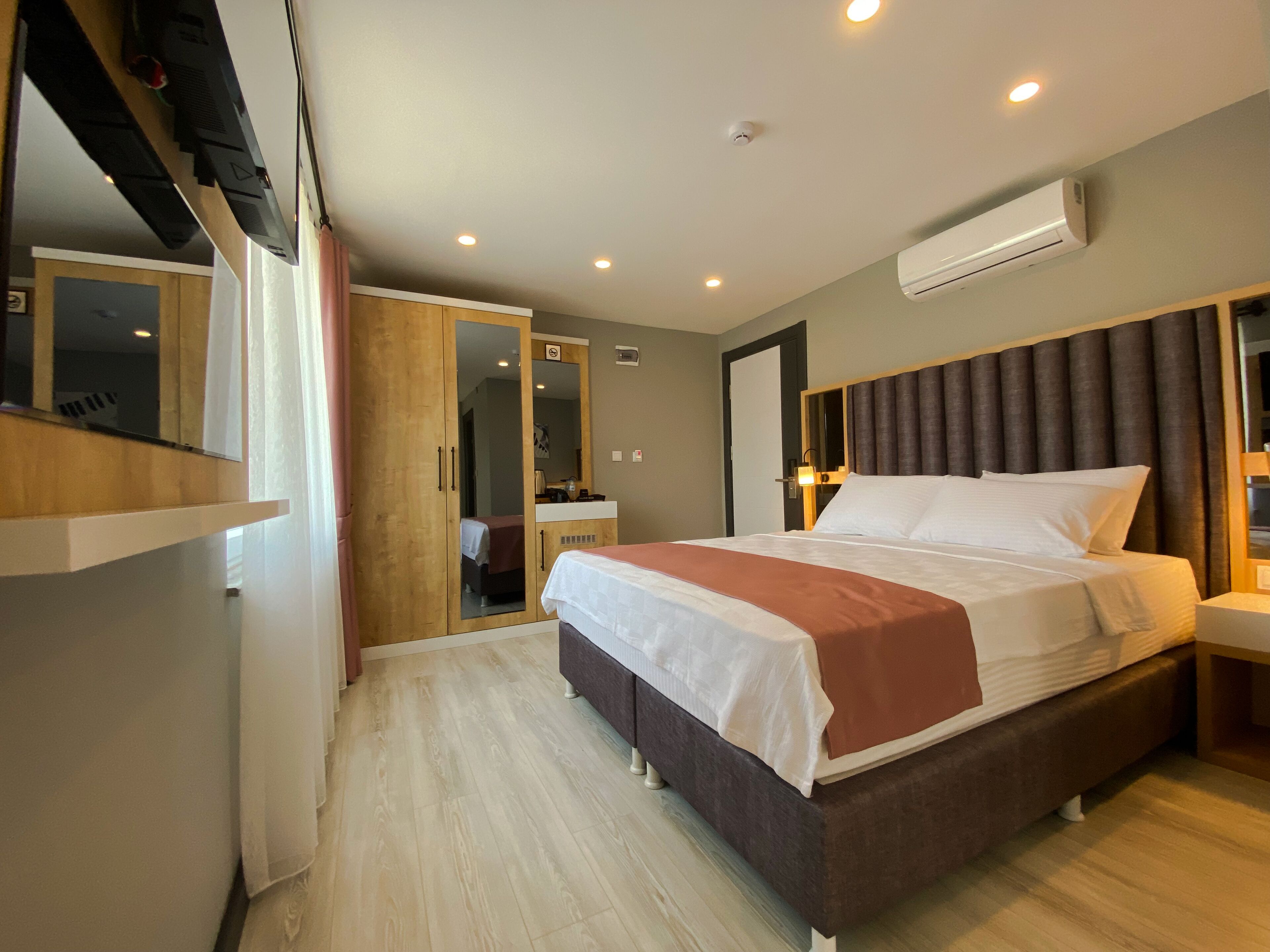 A modern hotel room with a large bed, a wooden wardrobe, a bedside table, an air conditioning unit, and a mirror.