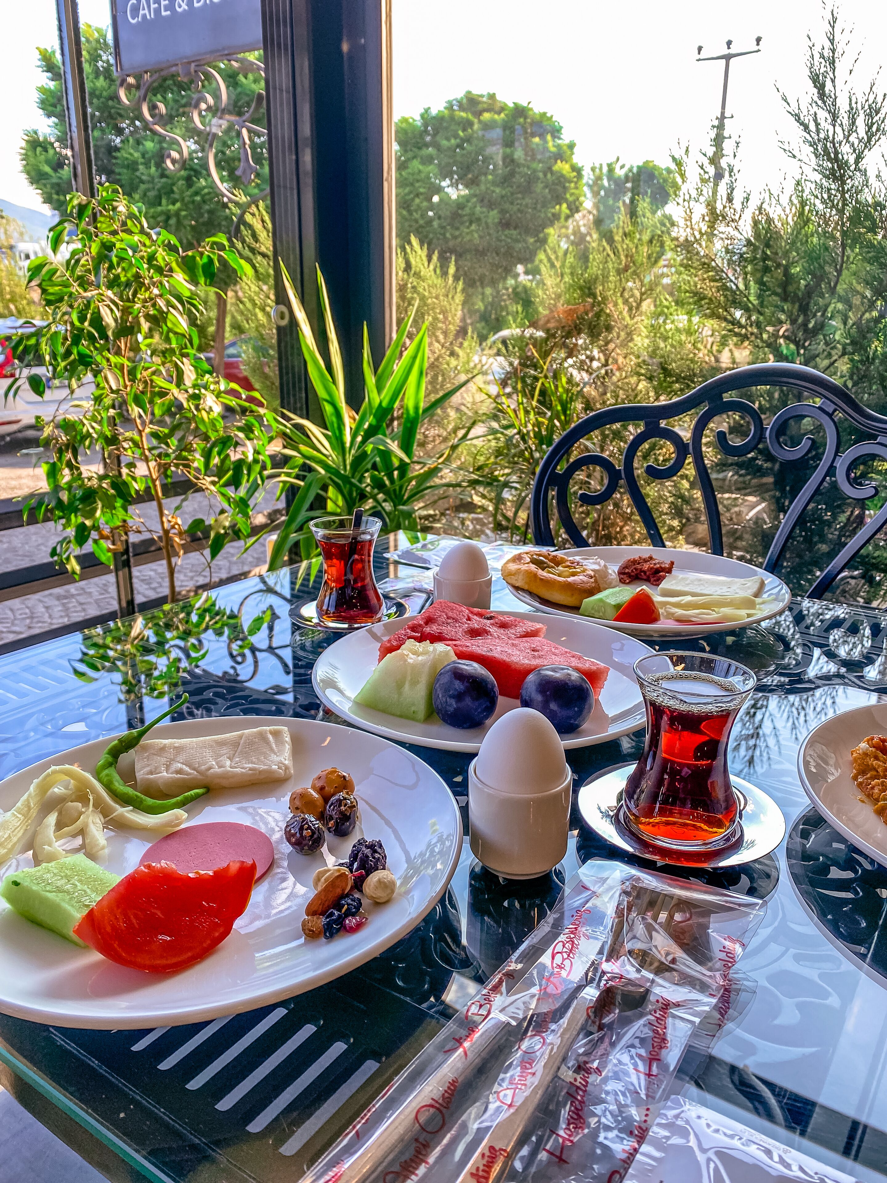 A table set with various dishes, including watermelon, eggs, and pastries, at a cafe with a view of greenery and parked cars.