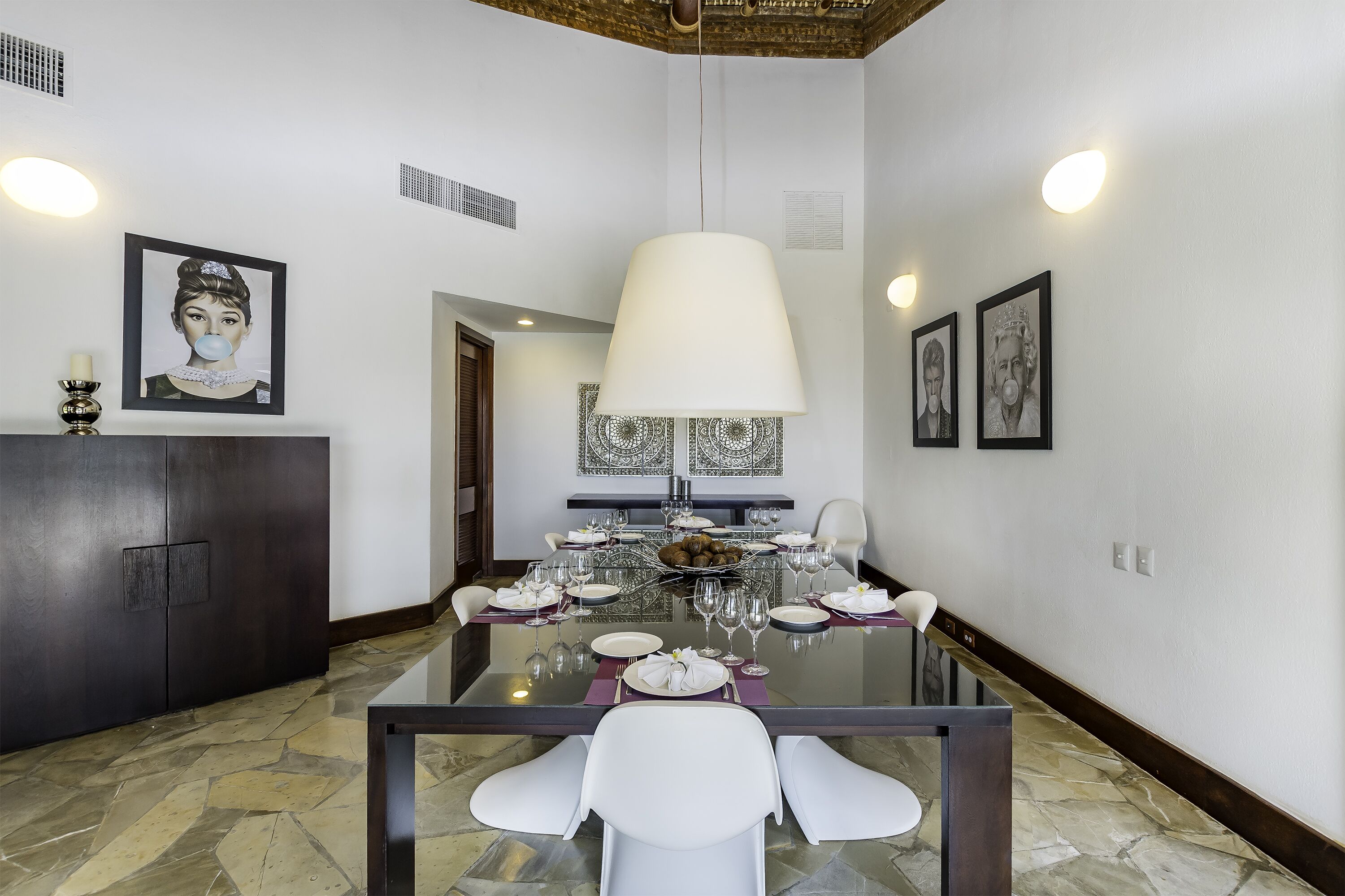 A dining area with a glass table set for six, white chairs, a dark wooden cabinet, and framed artwork on the walls.
