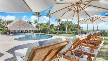 A pool area with lounge chairs and umbrellas, a thatched-roof structure, and palm trees.