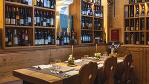 Restaurante com mesas e cadeiras de madeira, garrafas de vinho em prateleiras e um suporte para menu.