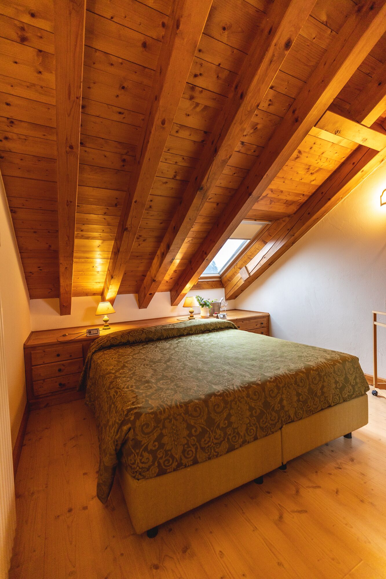 A bedroom with a sloped ceiling, wooden beams, a bed with a patterned bedspread, a nightstand, a lamp, and a small table with a plant.