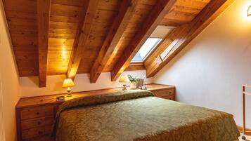 A bedroom with a sloped ceiling, wooden beams, a bed with a patterned bedspread, a nightstand, a lamp, and a small table with a plant.