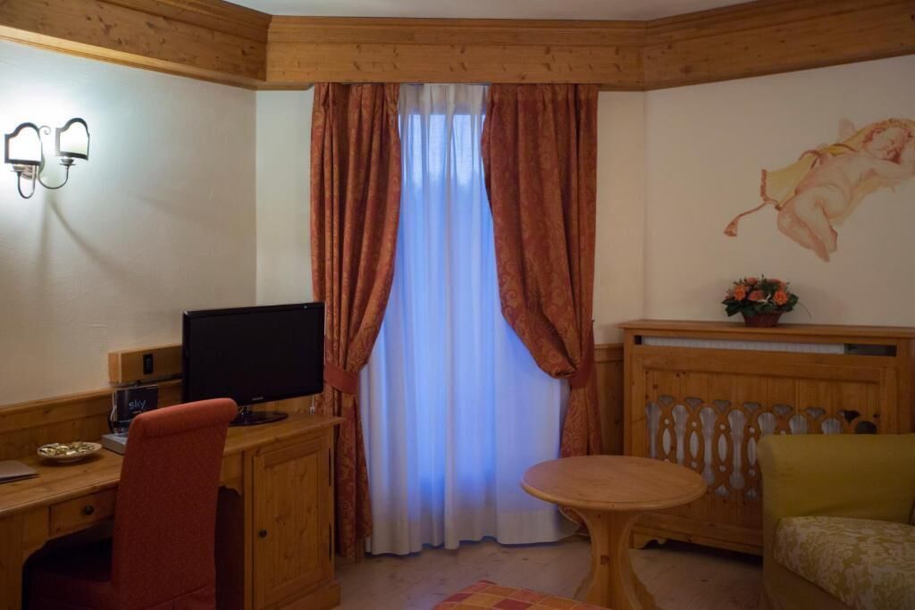 A room with a wooden desk, a computer, a chair, a round table, a sofa, a window with curtains, and a wall-mounted light fixture.