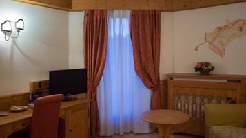 A room with a wooden desk, a computer, a chair, a round table, a sofa, a window with curtains, and a wall-mounted light fixture.