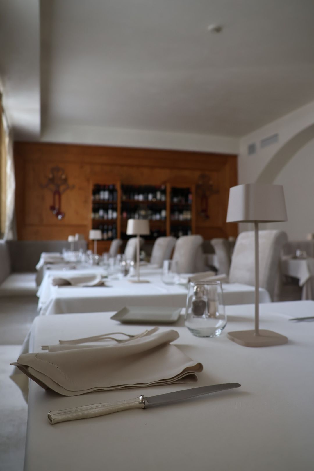 Ein Restaurant mit einem langen Esstisch, einem Weinschränkchen im Hintergrund und einer Lampe auf dem Tisch.