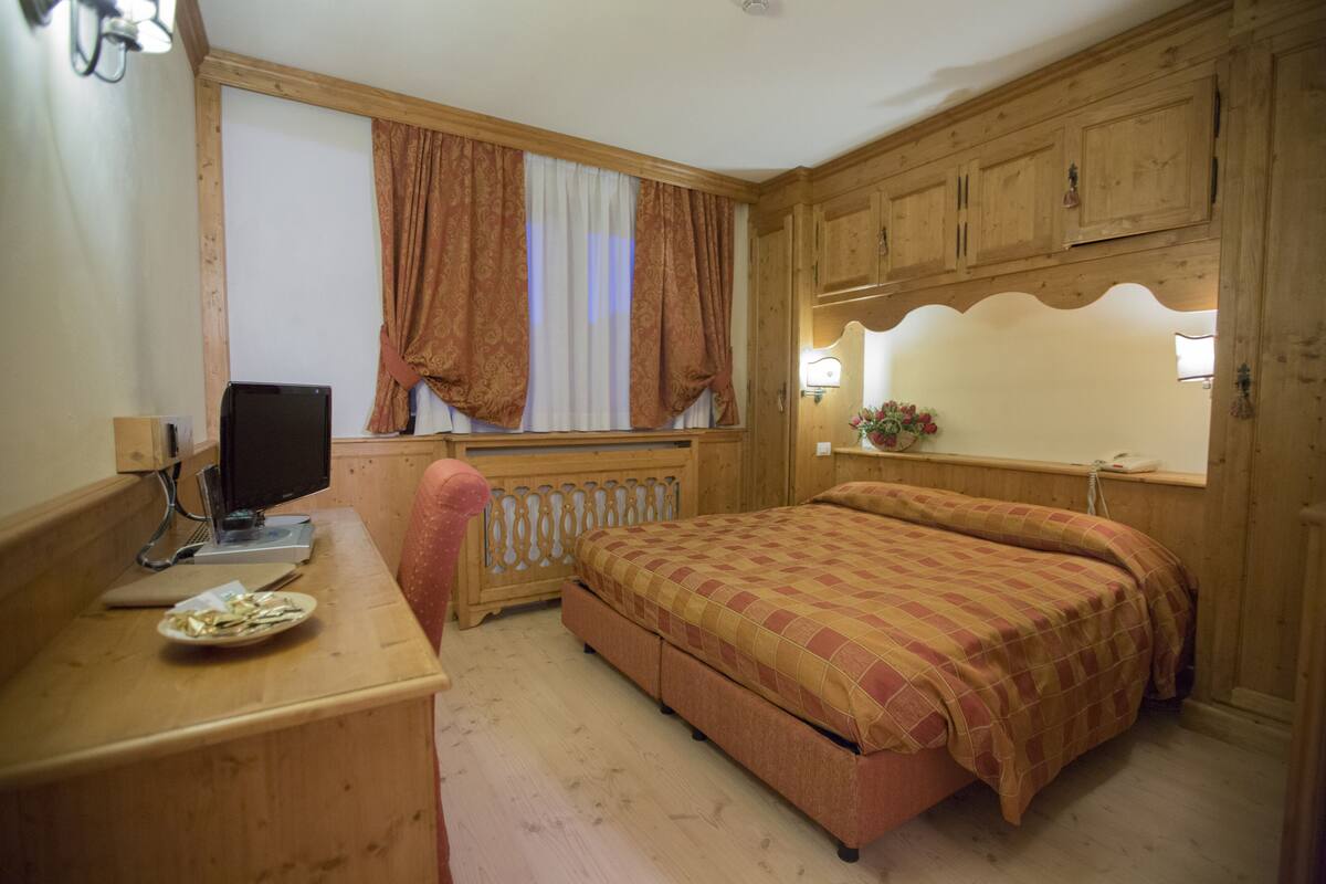 A wooden bedroom with a bed, a desk, a chair, a television, and a window with curtains.