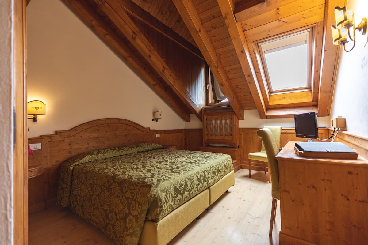 A bedroom with a wooden ceiling, a bed with a patterned bedspread, a desk with a computer, and a window with a view.