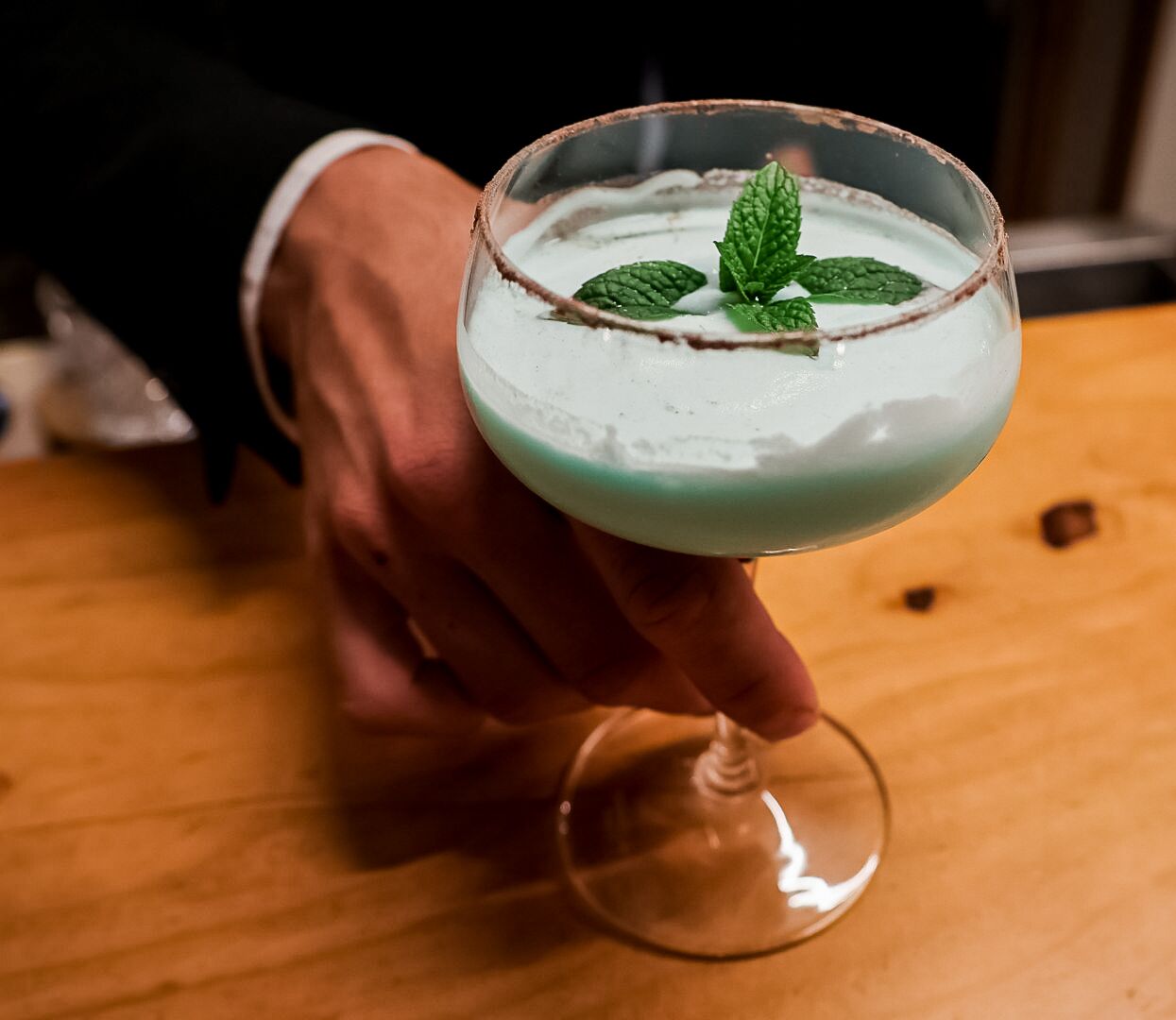 En person holder et cocktailglas med pynt af mynte på et træbord.