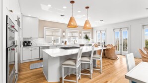 A modern kitchen with white cabinetry, a large island, and a dining area with wicker chairs. - Luxury Renovation in Seaside w/Expansive Rooftop Deck, Gulf Views, & 3 Kings (Santa Rosa Beach)