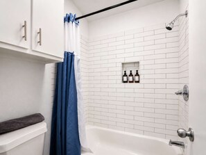 A bathroom with a white bathtub, a shower, a blue shower curtain, and a shelf with three bottles.