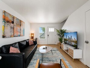 Ein modernes Wohnzimmer mit einem schwarzen Ledersofa, einem Couchtisch, einem Fernseher auf einem weißen Standfuß und einem großen abstrakten Gemälde an der Wand.