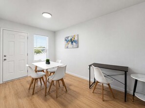 A modern dining area with a white table and chairs, a dark desk, and a painting on the wall.