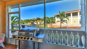 En balkon med grill, en fletstol og udsigt til en bygning med balkon og palmer.