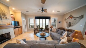 A spacious living room with a grey sofa, a coffee table, and a TV. There is a fireplace, a ceiling fan, and a view of the beach through large windows. - BEACHFRONT, 5BR/5BA, Sleeps up to 12 w/ Elevator & Pool! (St. George Island)