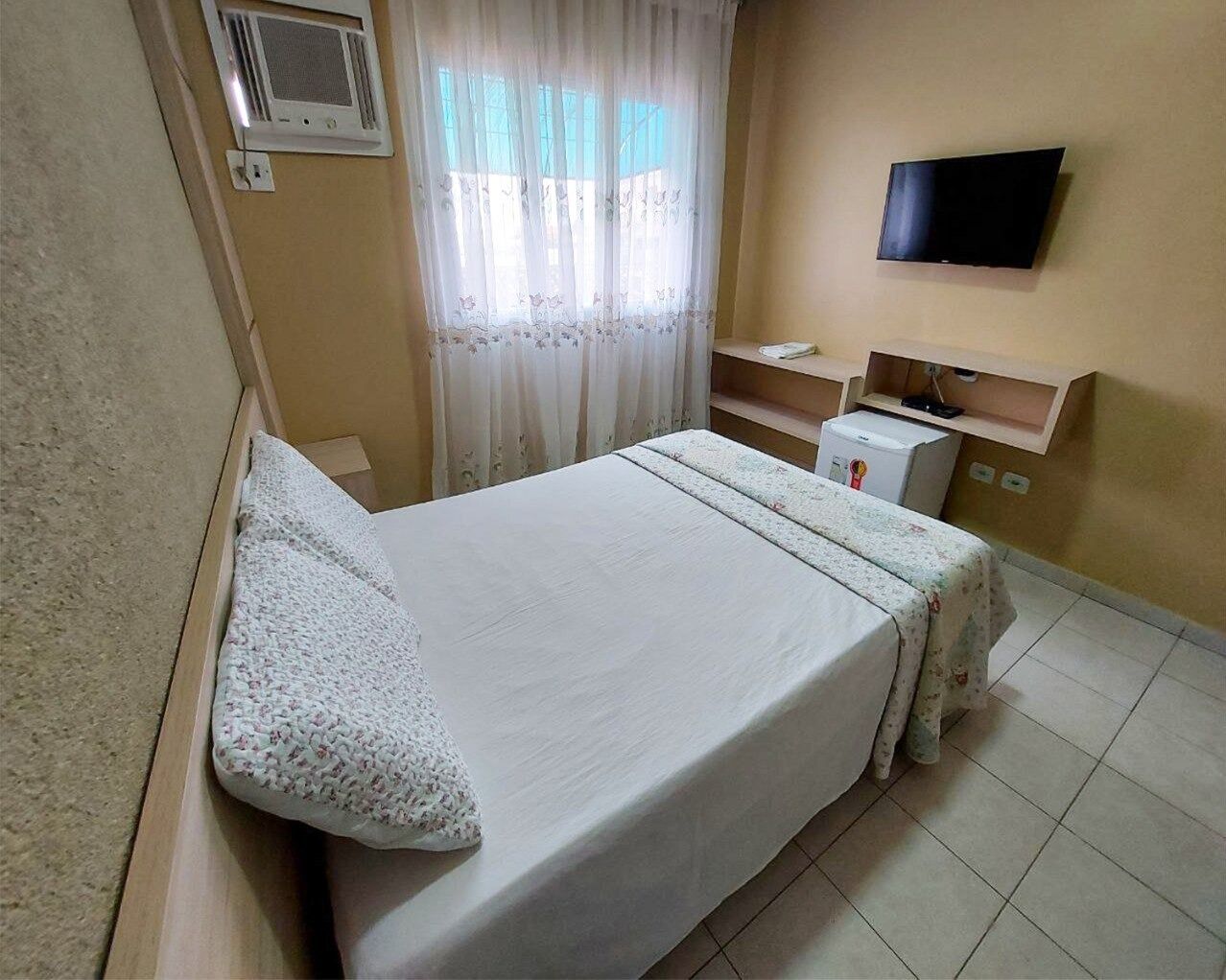 A hotel room with a bed, a television on the wall, a small desk, and a window with curtains.
