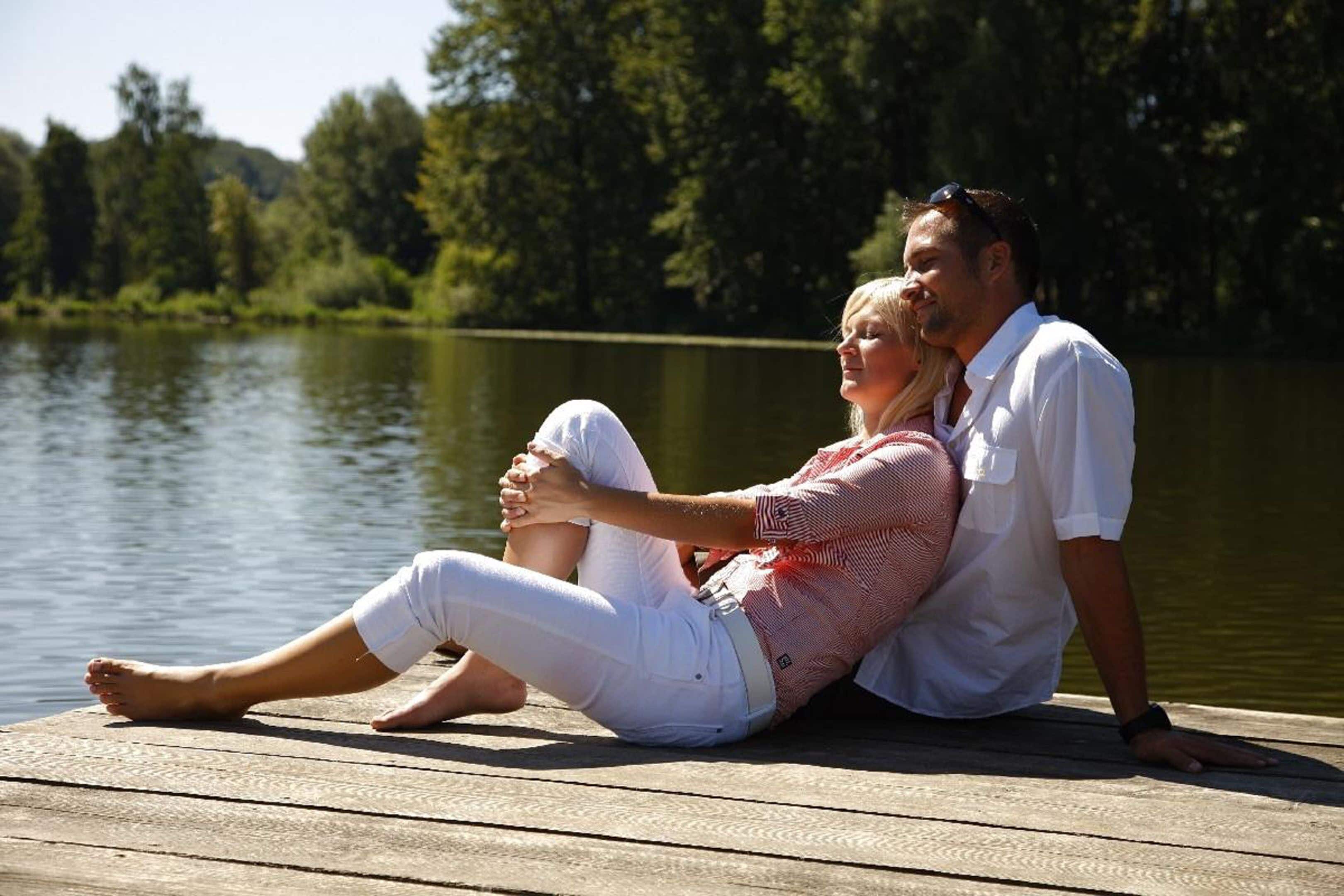 Una pareja sentada en un muelle de madera junto a un lago, con árboles al fondo.