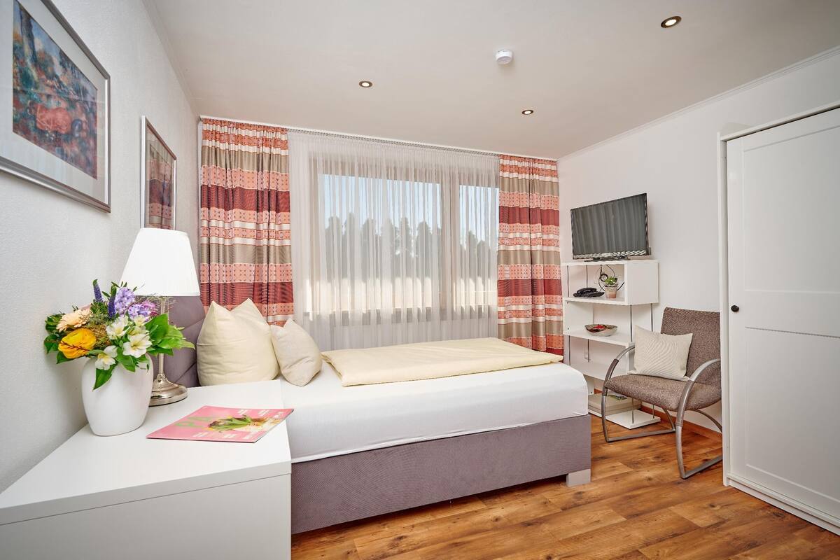 Une chambre à coucher avec un lit, une chaise, un téléviseur, un bureau avec une lampe et des fleurs, et une fenêtre avec des rideaux.