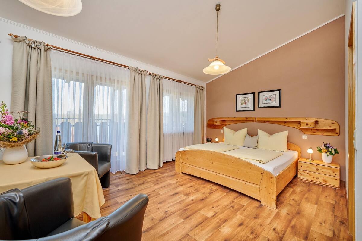 Une chambre à coucher avec un lit en bois, une table à manger avec des chaises, un vase de fleurs et une fenêtre avec des rideaux.