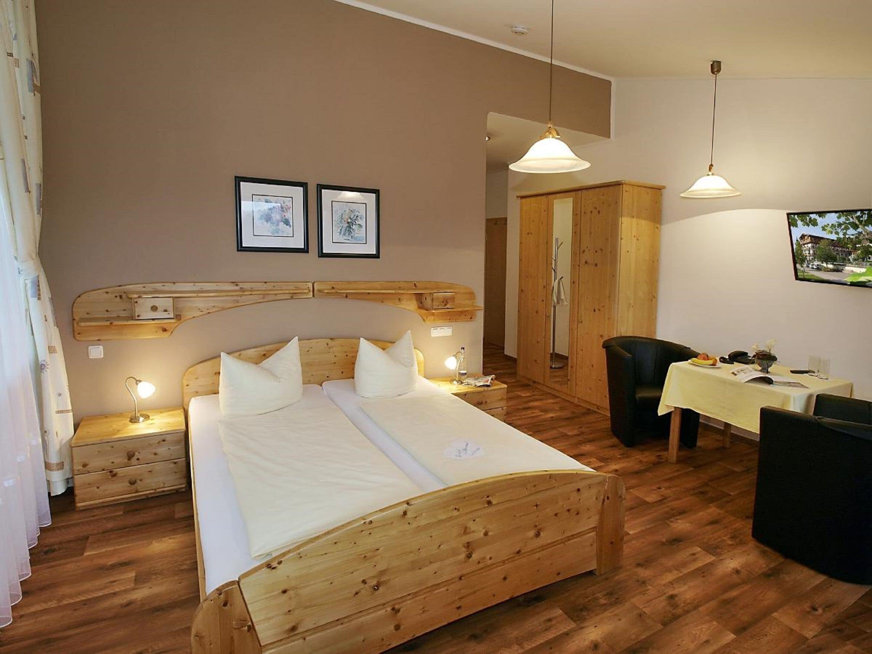 A wooden bed with two pillows, a bedside table with a lamp, and a small table with a chair in a room with wooden flooring and a wardrobe.