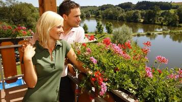 A couple standing on a balcony overlooking a lake with lush greenery and flowers.