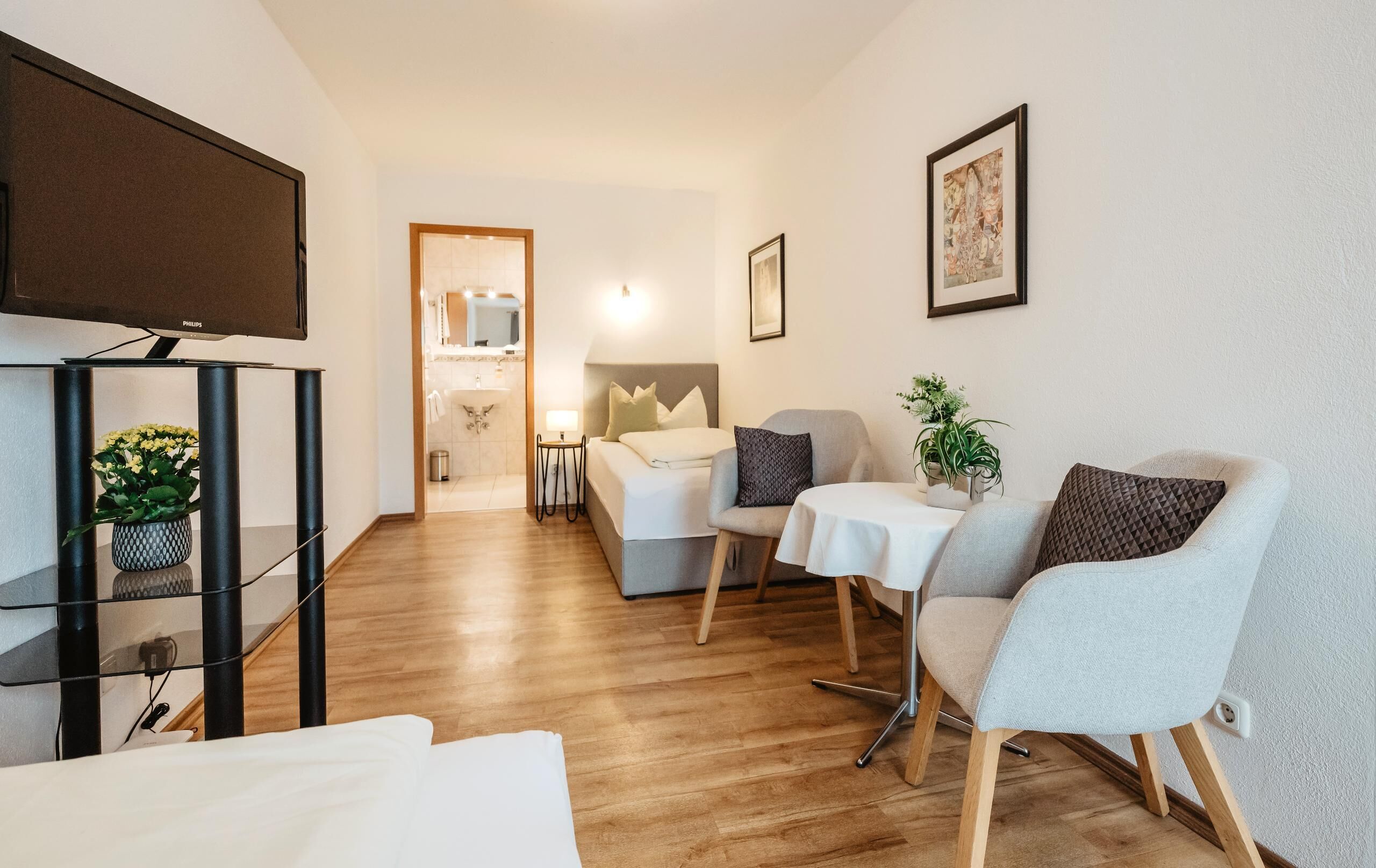 A modern hotel room with a wooden floor, a bed with white linens, a grey armchair, a small table with a white tablecloth, and a wall-mounted TV.
