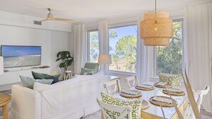 Una sala moderna con un sofá blanco, una mesa de comedor con individuales tejidos, un ventanal con vista a árboles y un ventilador de techo.