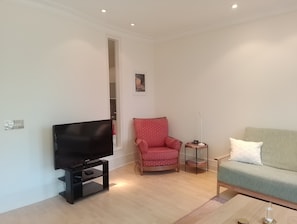 Un salon avec un téléviseur à écran plat posé sur un meuble, un fauteuil rouge à motifs, un canapé vert et une petite table d’appoint avec une lampe.