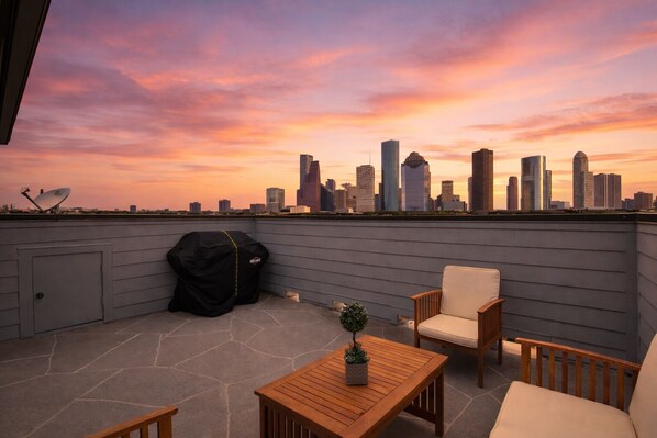A rooftop terrace with a city skyline view, a barbecue grill, and outdoor furniture.