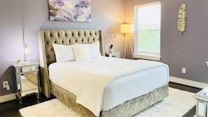 A bedroom with a tufted headboard, a large window with blinds, a bedside lamp, and a decorative feather wall piece.