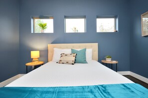 A bedroom with a large bed, three windows, a bedside table with a lamp, and a small plant.