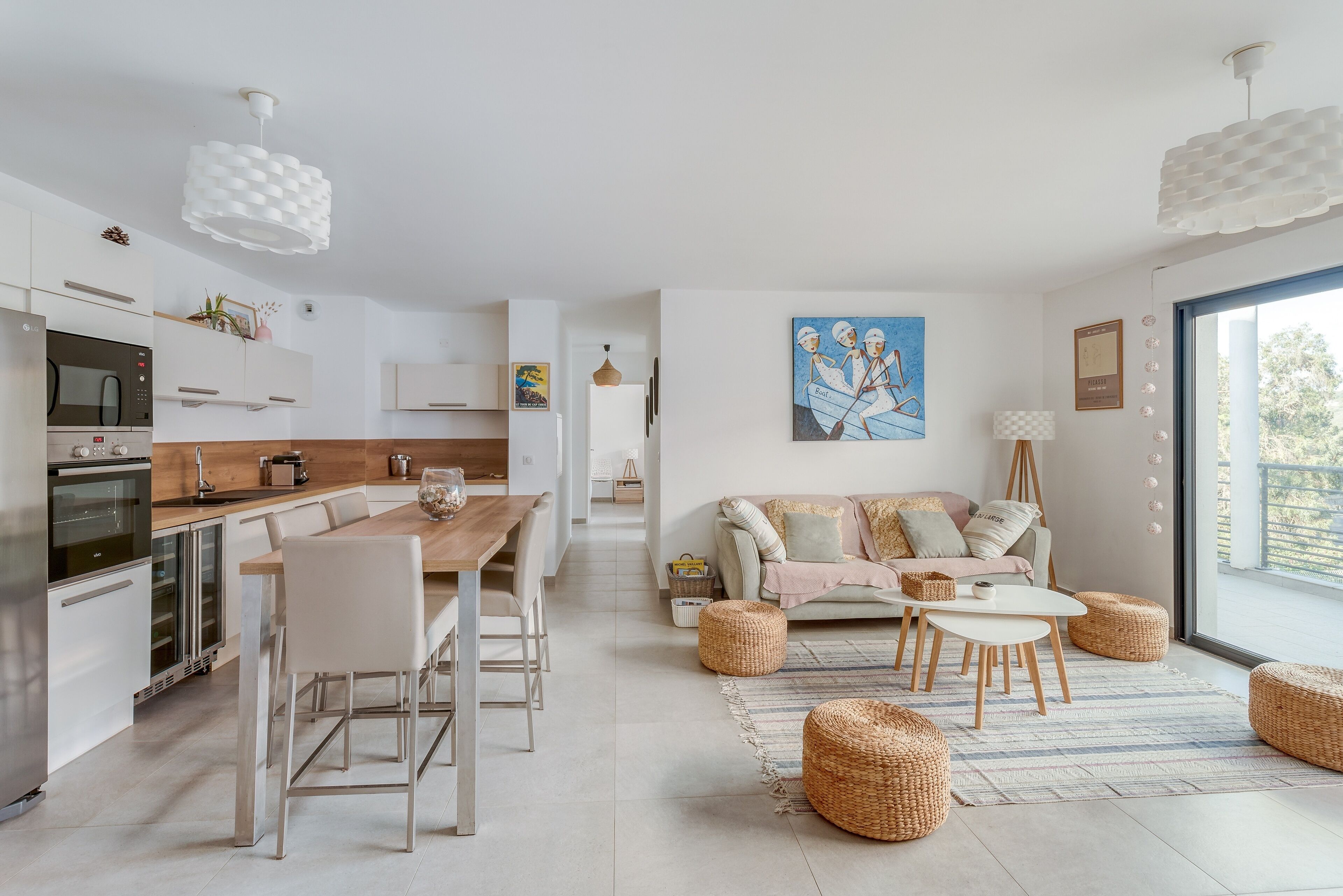 Une cuisine moderne avec des appareils électroménagers blancs, un coin repas avec une table et des chaises en bois, et un salon avec un canapé et des poufs.