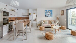 Una cocina moderna con electrodomésticos blancos, un comedor con mesa y sillas de madera, y una sala de estar con sofá y otomanas.