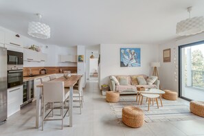 A modern kitchen with white appliances, a dining area with a wooden table and chairs, and a living room with a sofa and ottomans.