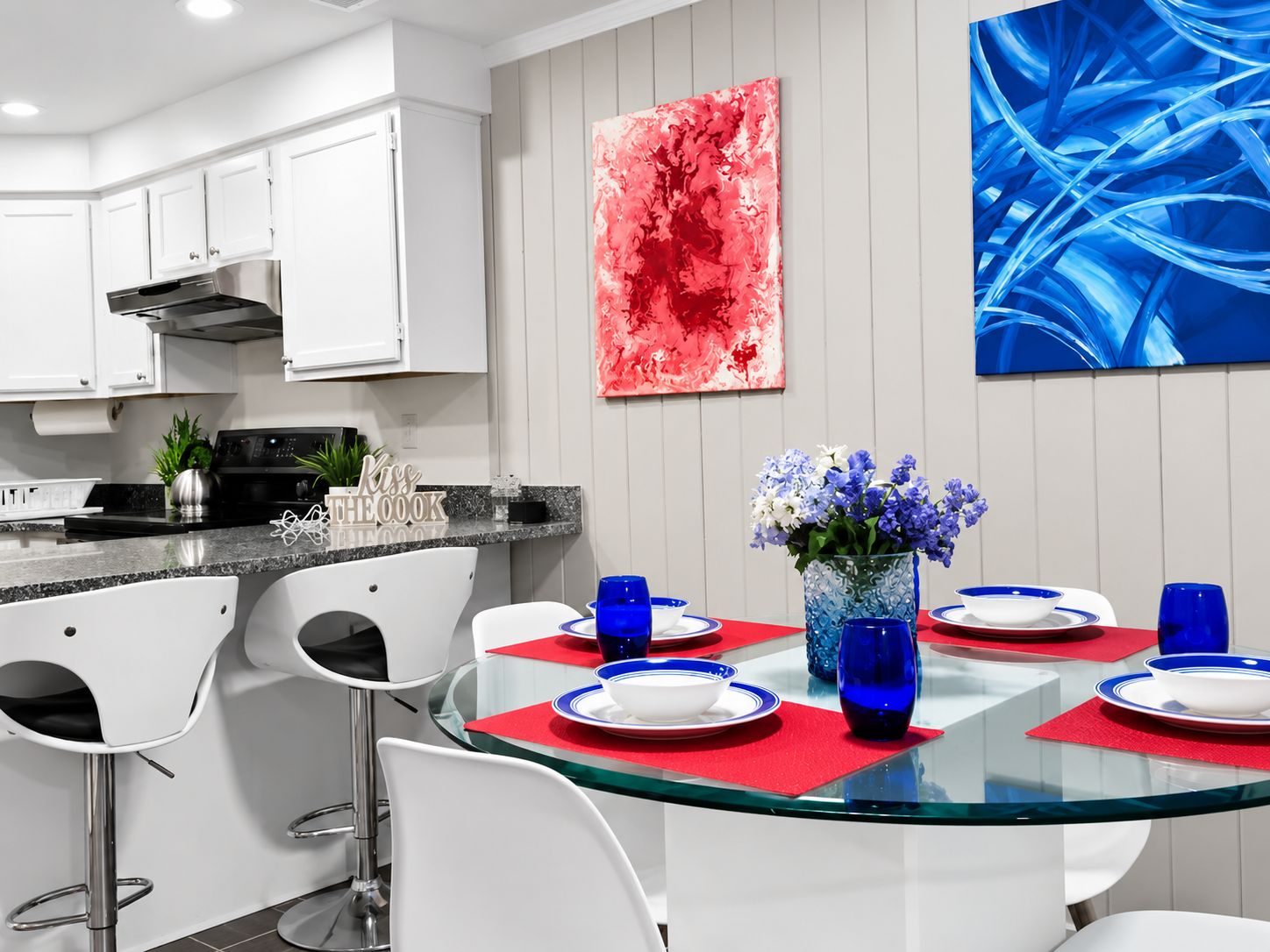 Una cocina moderna con armarios blancos, una estufa de acero inoxidable y un comedor con mesa de vidrio rodeada de sillas blancas.