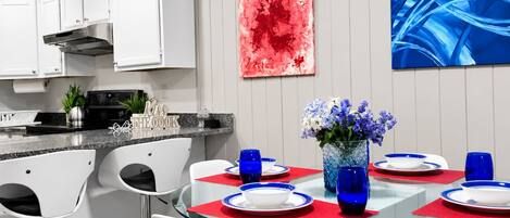 Una cocina moderna con armarios blancos, una estufa de acero inoxidable y un comedor con mesa de vidrio rodeada de sillas blancas.