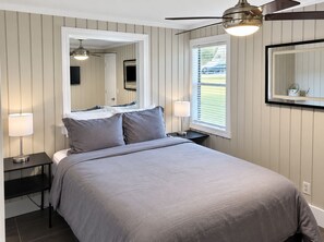 A bedroom with a bed, bedside lamps, a ceiling fan, a window with blinds, and a wall-mounted mirror.