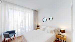 Quarto de hotel moderno com uma cama grande, uma cadeira, duas mesas de cabeceira com luminárias e uma janela ampla com cortinas leves.